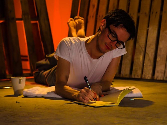 Nicholas Ryan lying on a concrete floor with pallets behind him and a coffee cup next to him writing narratives in a notebook