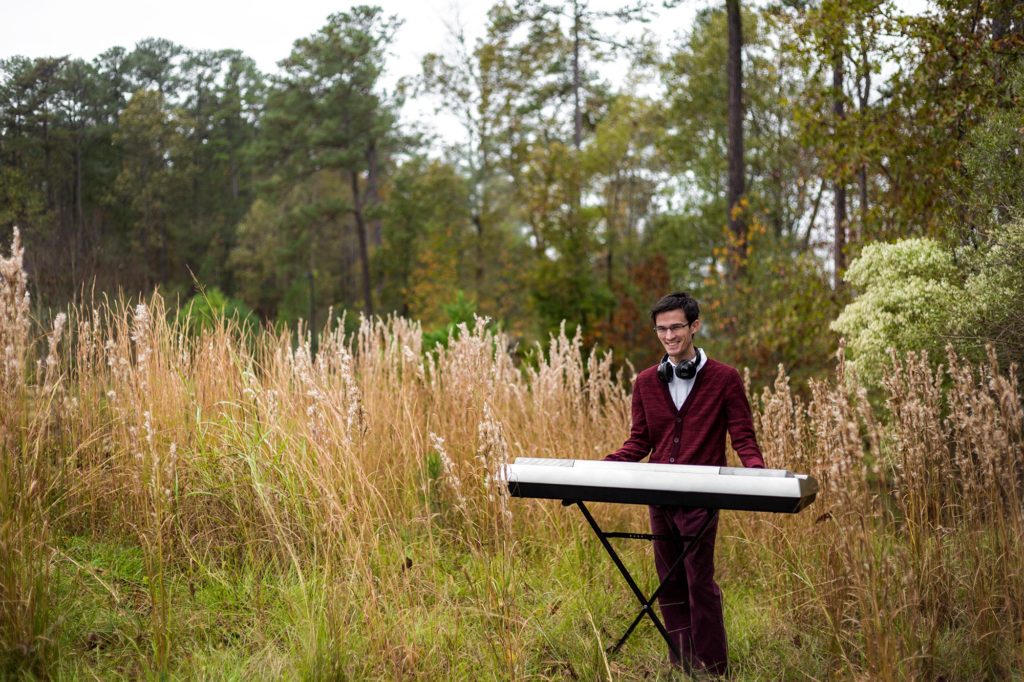 Nicholas Ryan wearing a red sweater playing music on an electric piano in a field of wheat