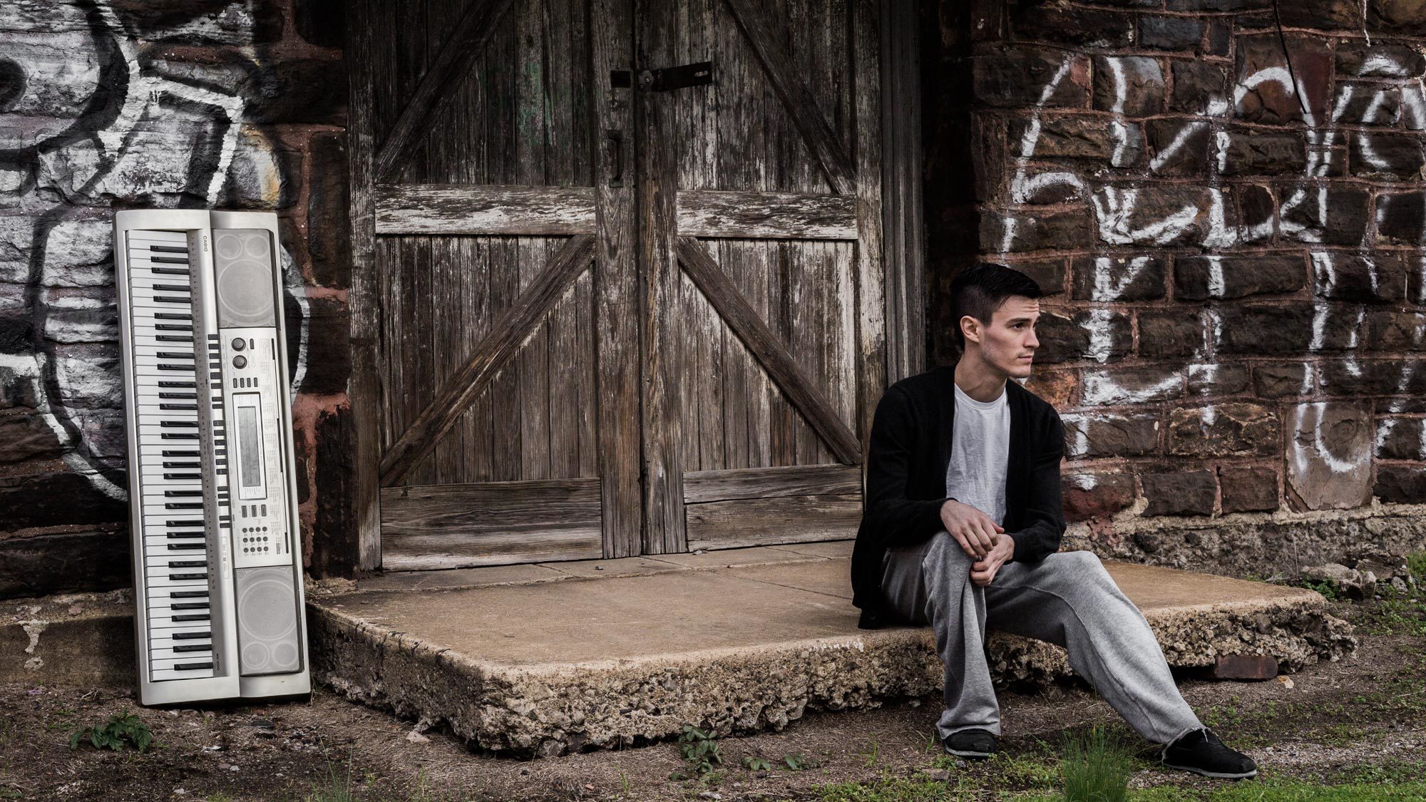 Nicholas Ryan sitting on a concrete slab in front of a distressed door with a an electric piano for music production to the side